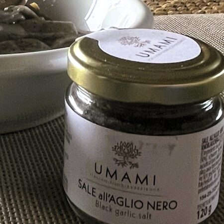 Vasetto UMAMI di sale all’aglio nero accanto a un piatto di pizzoccheri su tovaglietta intrecciata