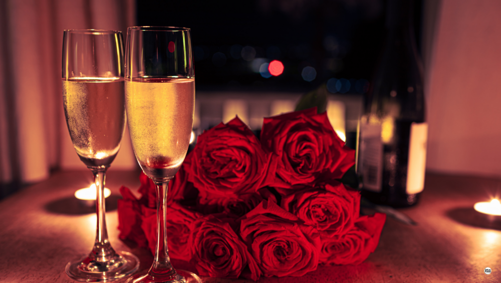 Tavola apparecchiata per una cena romantica con calici di bollicine, rose rosse e candele accese.