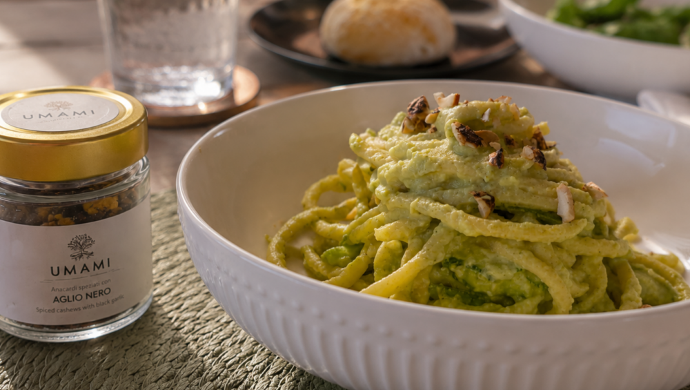 Piatto di bucatini con pesto di zucchine e avocado, anacardi tritati e vasetto UMAMI Anacardi Speziati all'Aglio Nero in primo piano