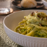 Piatto di bucatini con pesto di zucchine e avocado, anacardi tritati e vasetto UMAMI Anacardi Speziati all'Aglio Nero in primo piano