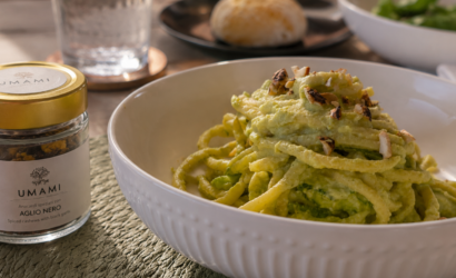 Piatto di bucatini con pesto di zucchine e avocado, anacardi tritati e vasetto UMAMI Anacardi Speziati all'Aglio Nero in primo piano