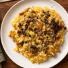 Risotto alla zucca con funghi, polvere di aglio nero e cacao amaro, servito in un piatto bianco su una tavola in legno.