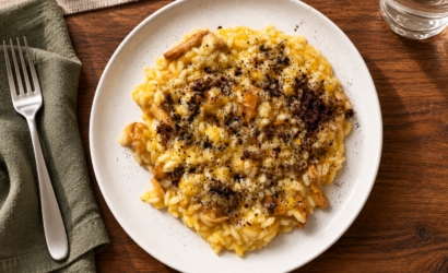 Risotto alla zucca con funghi, polvere di aglio nero e cacao amaro, servito in un piatto bianco su una tavola in legno.